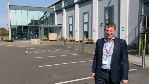 Matthew Burgess, Principal and CEO, wearing a light blue shirt and a navy suit with a red lanyard, standing outside the new Gloucestershire university campus. It is a large rectangular building with a glass entrance and car parking spaces outside.
