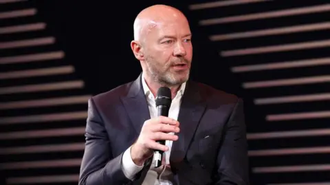 Getty Images Alan Shearer wearing a black suit and white shirt, talking into a microphone