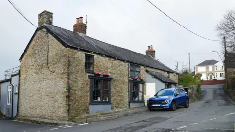 BBC An old brick pub with black window sills and two large windows on the ground floor. Outside a modern blue car is parked on the road, with faded road markings on the ground.