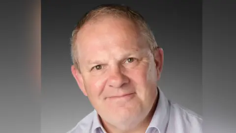 Royal Berkshire NHS Trust Steve McManus sitting in front of a grey background. He is wearing an open necked shirt and is smiling for the camera. 