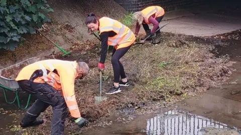 Volunteers in high viz on the floor of a river with almost no water lifting soil and silt