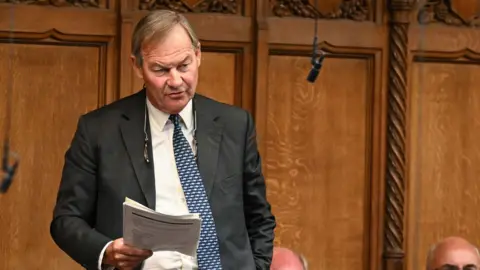 Rupert Lowe stands in the House of Commons, holding a document as he speaks. He is surrounded by seated members of Parliament. The chamber features traditional green benches and ornate wooden paneling.