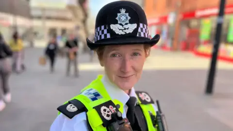 Woman in high-vis police vest and a police hat. She is stood on a street looking at the camera.