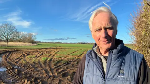 An older man with grey hair wearing blue gilet and black jumper. Behind him is a field with tyre marks as if a vehicle has recently driven there and a blue sky