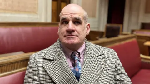 Leo looking at the camera. He is wearing a brown and cream checked jacket, a pink shirt and a blue patterned tie. He is sitting on a red bench.