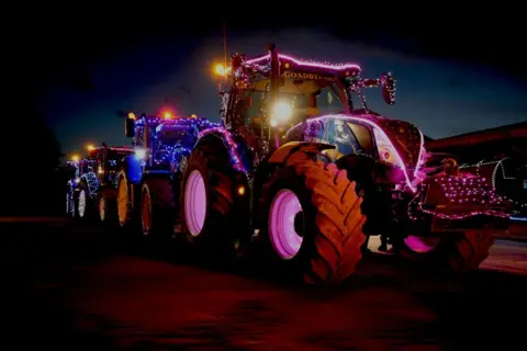 Martin Brandreth A line of tractors, each covered in lights and decorations. The photograph is taken at nighttime, and there are various different coloured lights covering the vehicles.
