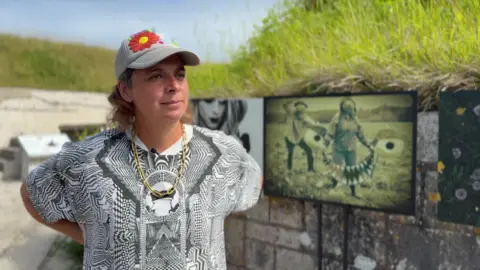 Rocca Holly-Nambi is standing inside the battery. They are wearing a gold chain around their neck and a cap with flower motif sewed onto the front of it.