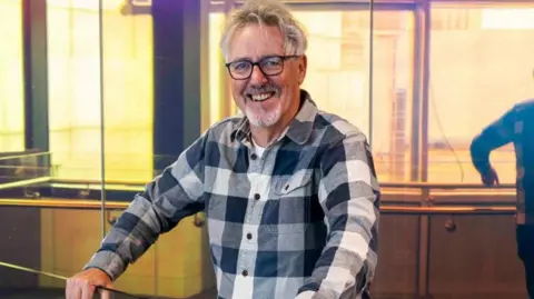 Griff Rhys Jones, a man with white hair and glasses smiling at the camera for a photo inside a studio. He is wearing a long sleeved checkered shirt.