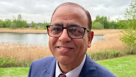 Sam Read/BBC Mohammad Yasin with short dark hair, wearing glasses and smiling at the camera. He has a blue jacket over a pink shirt and purple tie. He is standing on grass with reeds and a river behind.