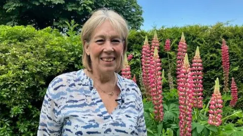 Lynda Sawyer smiling into the camera arsenic  she stands successful  beforehand   of a precocious   greenish  hedge and gangly  pinkish  flowers. She has blonde hairsbreadth  chopped  into a bob and is wearing a bluish  and achromatic  patterned blouse.