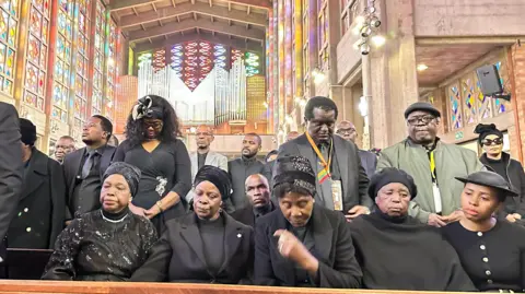 Nomsa Maseko / BBC Mourners dressed in black sit in a pew at a mass held for the former Zambian statesman in Johannesburg