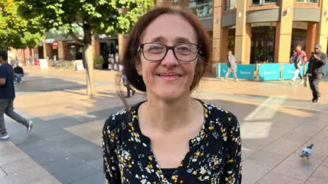 Joy Adams is pictured in Broadgate, in Coventry city centre, she is smiling at the camera and has glasses and is wearing a floral print blouse