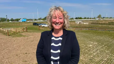 A woman with curly blonde hair, a blue top and blazer, stood in front of a field and road.