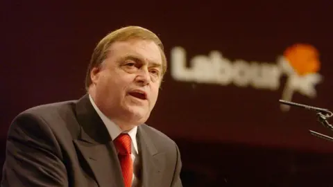 BBC John Prescott wearing a suit and red tie with a Labour sign in the background