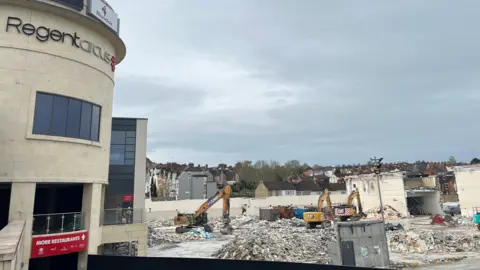 The Regent Circus site where the Morrisons supermarket used to be with three yellow diggers, rubble and the remains of some buildings in the background. Further into the background, there are houses and in the foreground you can see the remaining Nando's building and a large 'Regent Circus' sign. 