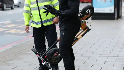 Anonymous shot of a person wearing a black tracksuit and black gloves carrying a folded e-scooter talking to a police officer who is wearing black trousers and a reflective jacket. They are stood on the pavement and the shot shows the middle of their bodies only.