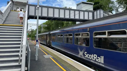 An image of a blue ScotRail train at a small passenger train station. The station looks new and clean. There is a staircase and lifts that connect the platforms above the train.