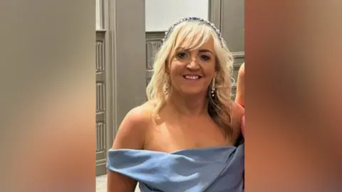 Michele Slane A woman is smiling into the camera. She's dressed in a light blue formal dress. She has blond hair, which is curled and she's wearing a hairband and dangling earrings. 