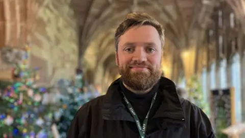 A man with blonde hair, a brown beard and a black top in a room with the blurred shapes of trees behind him