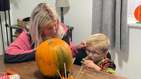 Noa sat at a table with a large pumkin in front of him, he's holding a marker pen to decorate the pumpkin, with his mother Sophie sat next to him, wearing a pink and black jacket