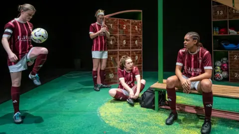 Nicola Young Four women in a red and white football kits are in a stage set up of a changing room. The woman on the far left is kicking a football up with her knee, another is standing by a locker, a third is sat cross-legged on the fourth and the woman on the right is sat on a bench