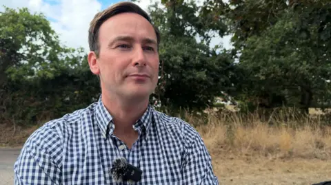 Tom is standing on grass with hedge behind him. He is wearing a blue and white checked shirt with a microphone attached to it