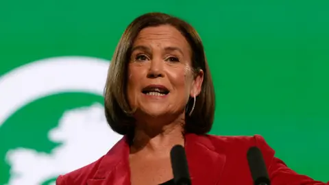 PA Media Mary Lou McDonald is pictured speaking at her party's annual conference. She has shoulder length brown hair and is dressed in a red blazer and back top, gesturing with her left hand. Behind her the backdrop is bright green and on the podium in front of her are the words 'Building Ireland's future'.