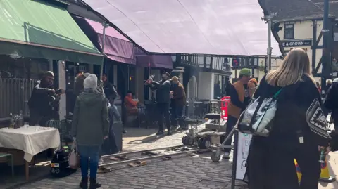 Holly Nichols/BBC A cobbled street in Hitchin where a film crew are filming a drama. On the right are various shop awnings. In the middle, an awning has been put up by the film crew on metal legs. There is a camera gantry on the floor with a seat on wheels. Men are milling around looking at lights or cameras. A woman in the foreground on the left with her back to us is wearing a hat, coat, jeans and boots and has a handbag in her left hand. On the right is a backview of another woman with blonde hair, wearing a black coat and a bag over her shoulder. 