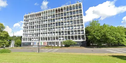 Google A high, brutalist building on the side of a main road. The sky is bright blue in the background and there are trees and grass around the building. There are several masts on top of the building. 