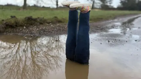 Sophie Montague/BBC A pair of fake legs are placed in a water-filled pothole with the feet sticking out at the top. The legs are constructed from blue jeans and there are colourful shoes on the fake feet. The pothole is large and is at the side of a road. The water is muddy