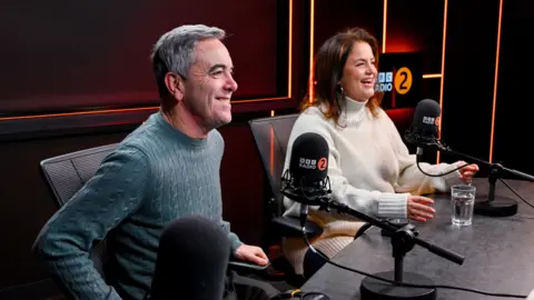 James Nesbitt and Ruth Jones laughing, sitting in front of Radio 2 mics. Nesbitt wears a blue knitted jumper, and Ruth Jones wears a while turtle neck