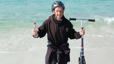 Bob Brown from Saltash, Cornwall stood on a beach in Australia having completed his challenge. He is stood just on the shoreline holding his scooter in one hand and a bottle of water in the other. He is wearing a black hoody and a black cycle helmet. 