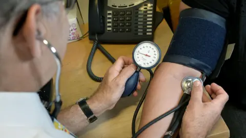 PA Media A doctor is using medical equipment including a blood monitor cuff to monitor someone's blood pressure. 