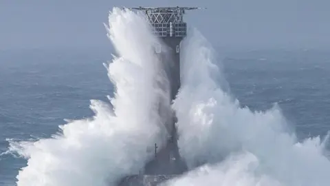 A massive wave crashing up against a tower in the sea. 