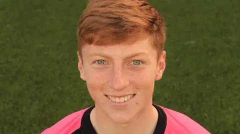 A young man with ginger hair in a pink football top with grass in the background