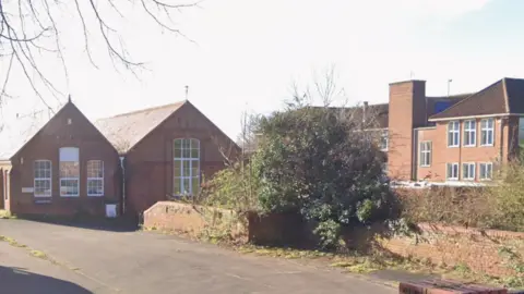 A Google street view image of the entrance to the car park of a former school. There are two building on the left of the image, a brick wall on which there is an overgrown bush in the middle of the image, and then a large building on the right of the image behind the bush.