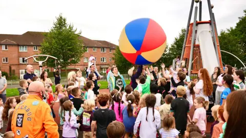 a group of young people in a play park chase a giant multicoloured ball