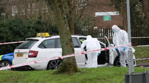 Two men in white forensic suits examining a white taxi with its door open. In the foreground police tape is tried to a street light.