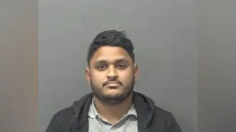 A mugshot of a man standing in front of a grey screen and wearing a hooded top.