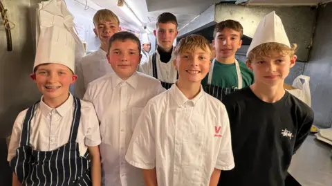 Seven teenagers, all boys, standing in a busy kitchen. All of them are wearing chef hats. The rest are wearing chef coats and aprons. 