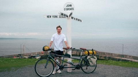 A man in a white tshirt and black shorts stood next to a tandem bicycle at the John O'Groats sign in Scotland
