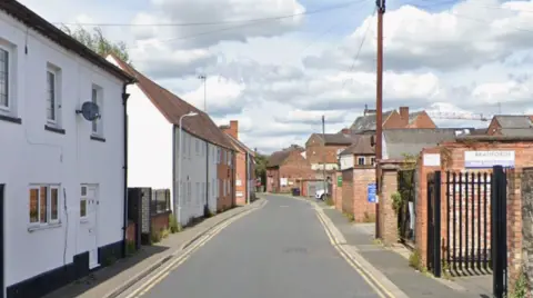 Google A generic photo of a street with buildings including white buildings on the left. Black gates are in the foreground on the right of the road and the street goes through the middle of the image.
