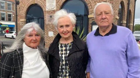 BBC Three Yarm Town councillors standing in a line outside a brick building. Pamela Smailes has long, grey-blonde hair and wears a high collared white blouse under a black jacket with a white check design. Barbara Wegg has short white hair and wears a black coat over a patterned top. Bob Wegg has short, receding white hair and wears a lilac jumper over a black shirt.