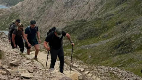Spinal Research Sam Baldwin walking up Snowdon using two black walking sticks. Behind him are family and friends.