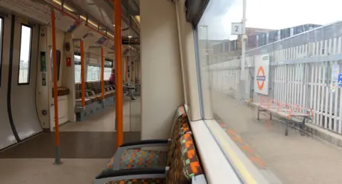 The inside of a London Windrush train showing a few seats and corridor, as well as an overground station outside the train window