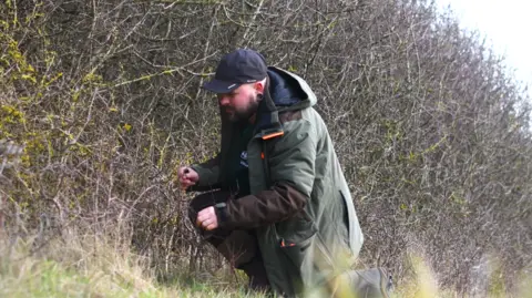 BBOWT A man in a large green coat and black baseball cap is down on one knee scouring a hedgerow for eggs.