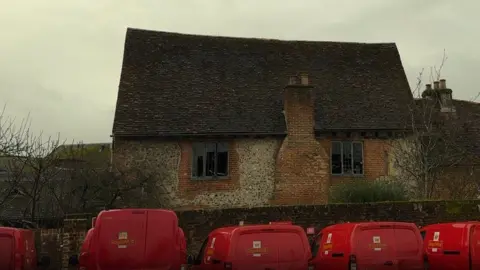 Line of red post office vans parked up in front of a brick wall where a brick and stone building stands behind the windows have been blown out