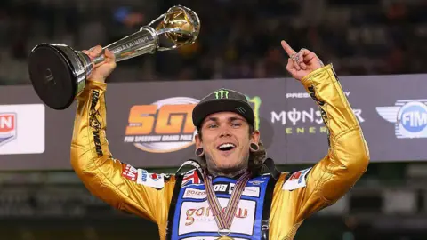A man wearing a black cap and a gold coloured overall stands with his hands in the air. He is holding a large gold trophy and has a medal around his neck.