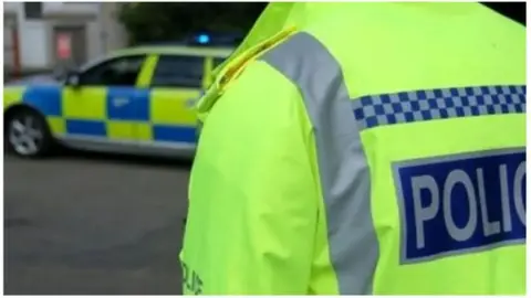BBC The torso of a police officer is wearing a bright yellow high vis jacket with the word "police" written on their back in blue and white writing. In the background is a police car