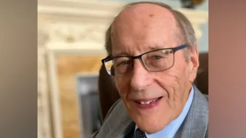 Supplied Barrie Tomlinson, side on, looking at the camera and smiling, wearing glasses, a suit and a tie and blue shirt. He is in front of a fireplace that is slightly blurred. 